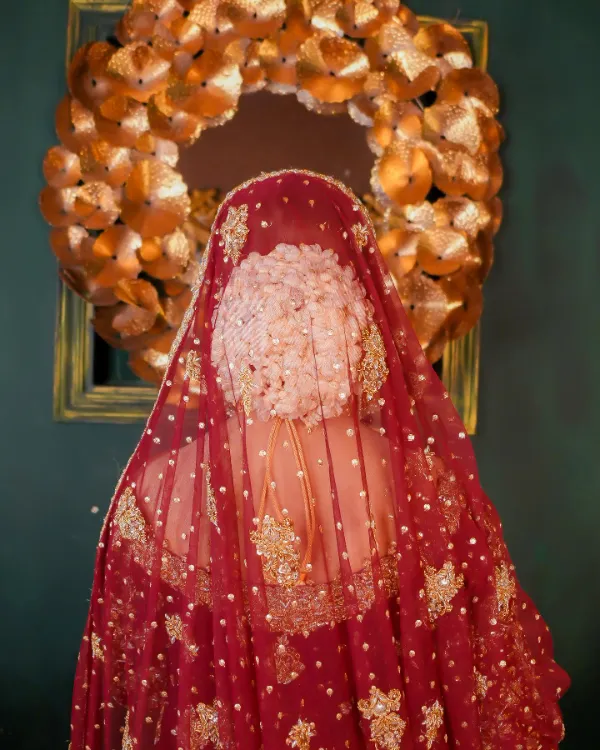 Bride Rabeeca Khan, back view, draped in a red and gold lehenga, facing a floral backdrop.