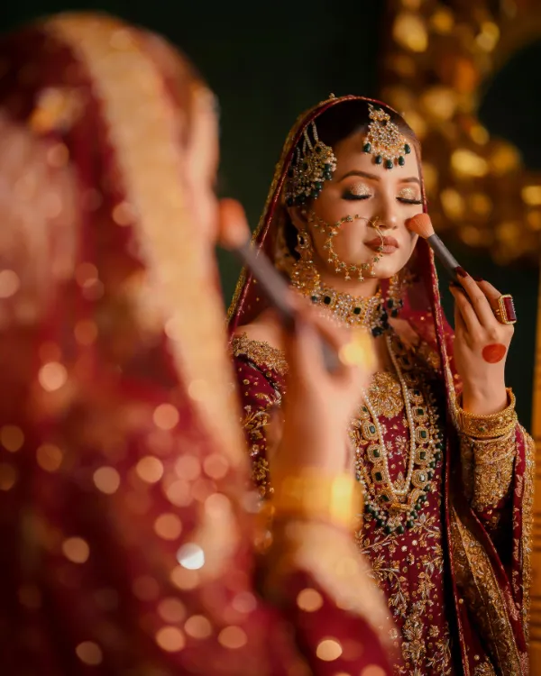 She dressed in a luxurious bridal outfit, applying makeup in front of a mirror, with a focus on her elegant makeup brush and traditional jewelry.